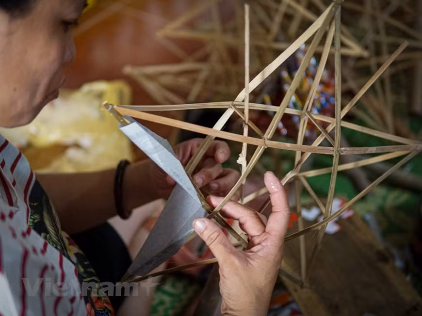 Selon Nguyen Thi Tuyen, de nombreuses étapes sont nécessaires pour perfectionner une lanterne. Le savoir-faire demande de la patience, de la minutie et de la concentration. L'étape la plus difficile consiste à fixer le cadre de la lanterne. La beauté du produit repose essentiellement sur ce cadre qui doit être équilibré et robuste. Il est ensuite recouvert de papiers glacés colorés qui sont découpés en six parties nécessaires d'une étoile. Chaque pièce est collée sur le cadre avec soin pour éviter tout décollement. À l'étape suivante, l'étoile est décorée de rubans et est entourée d'un fin cadre en bambou jaune. Une poignée en jute est enfin apposée pour tendre le cadre et mettre les lanternes à sécher. Photo: VietnamPlus