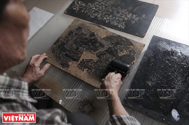 De l’encre noire est appliquée avec un rouleau sur la plaque de bois gravée, avant qu’une feuille de papier ne soit posée dessus.